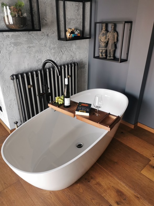 Wolnostojąca wanna z drewnianą półką, na której znajduje się butelka wina, kieliszek i książka, w tle szara ściana z dekoracyjnymi półkami i grzejnik retro.