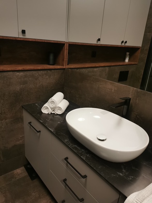 Nowoczesna łazienka z owalną umywalką nablatową, czarną armaturą i szarą szafką z podłużnymi uchwytami, uzupełniona o zwinięte białe ręczniki.