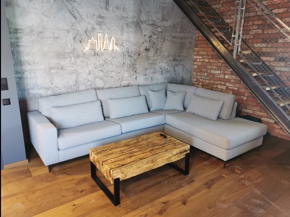 Szary narożnik z poduszkami, drewniany stolik kawowy z metalowymi nogami, betonowa ściana z dekoracją świetlną w kształcie panoramy miasta, schody metalowo-ceglane w tle