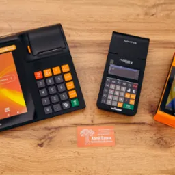 Trzy terminale płatnicze Novitus (Next Pro, Nano II online, One) w kolorach czarnym i pomarańczowym, ułożone obok wizytówki na drewnianym blacie.