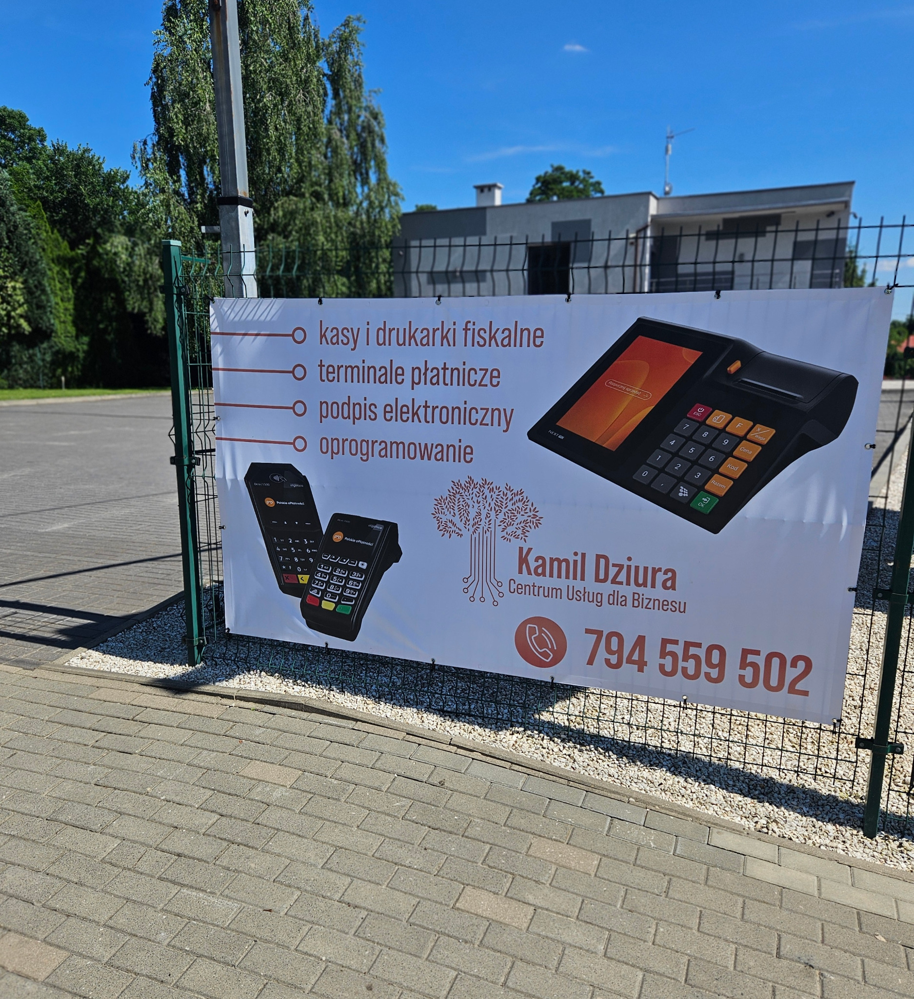 Baner reklamowy na ogrodzeniu, prezentujący kasy fiskalne, terminale płatnicze, podpisy elektroniczne i oprogramowanie, z danymi kontaktowymi Kamil Dziura Centrum Usług dla Biznesu, numer telefonu...