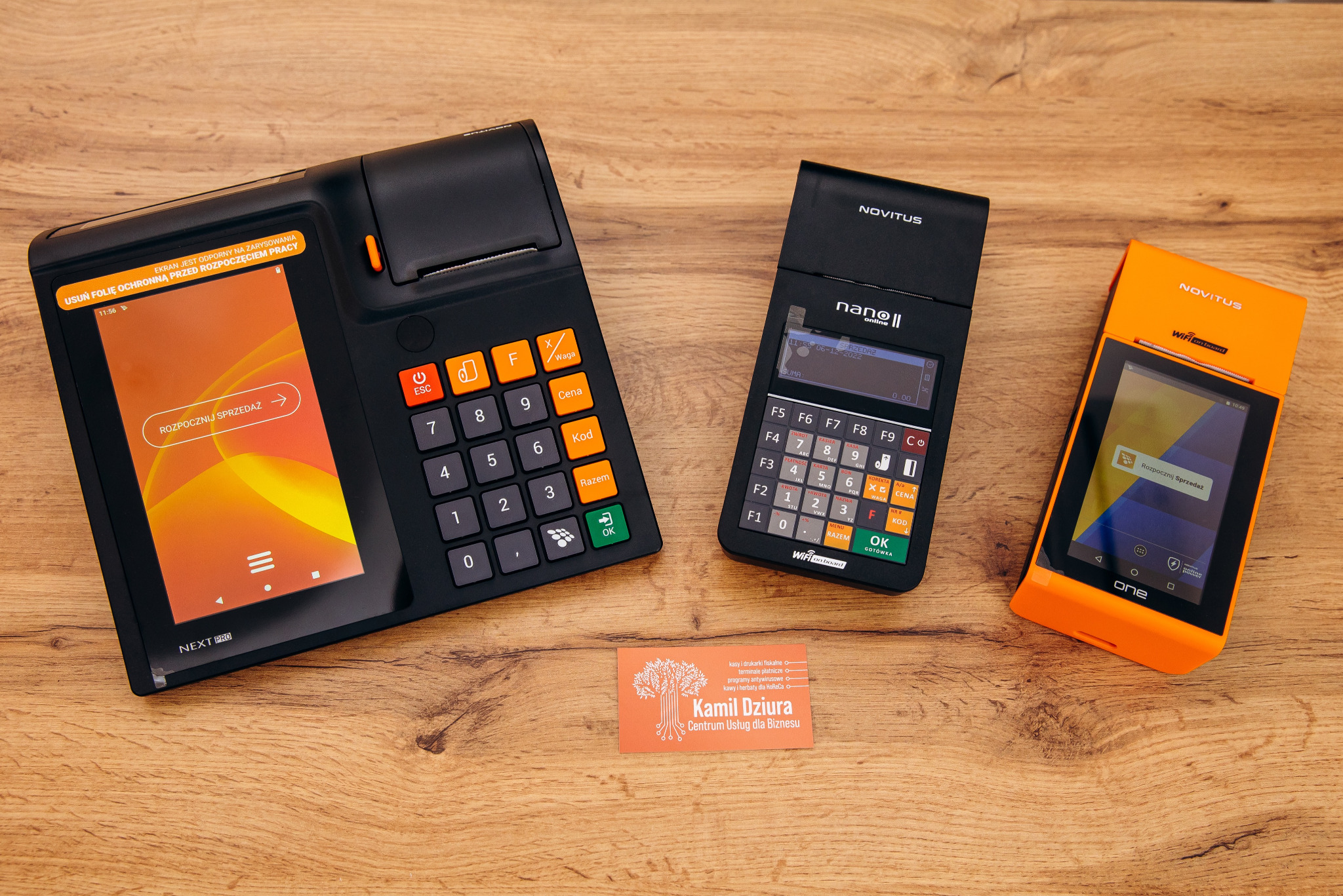 Trzy terminale płatnicze Novitus (Next Pro, Nano II online, One) w kolorach czarnym i pomarańczowym, ułożone obok wizytówki na drewnianym blacie.