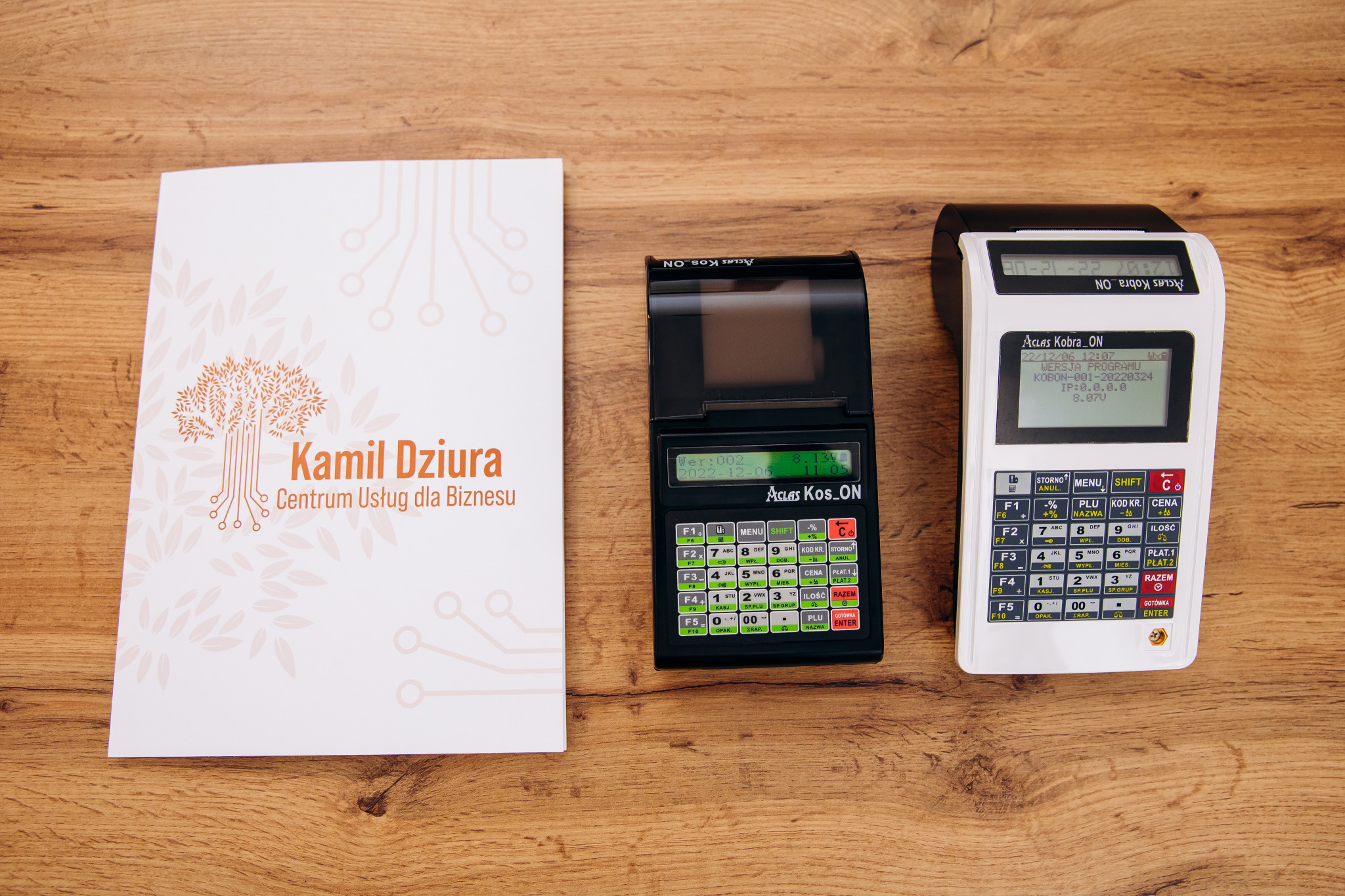 Dwa modele kas fiskalnych Aclas (Kobra i Kos_ON) obok ulotki firmowej z logo drzewa z elementami obwodu drukowanego, na drewnianym blacie.