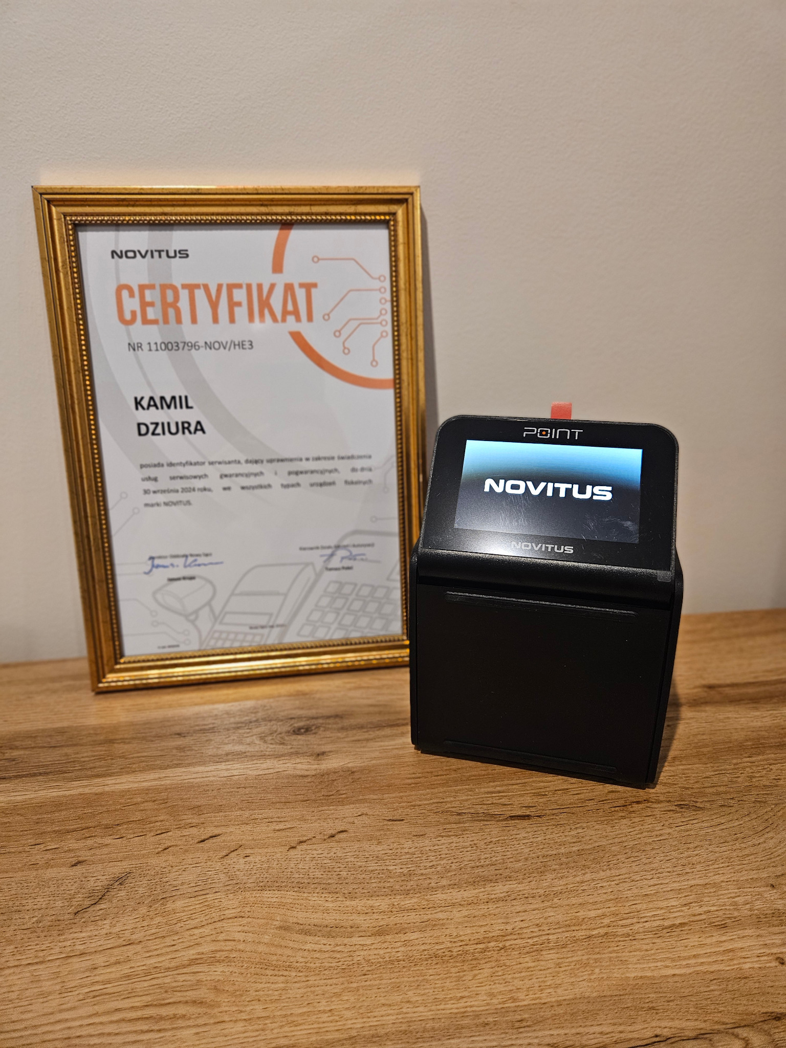 Certyfikat serwisowy Novitus w złotej ramce, obok czarna kasa fiskalna Novitus z wyświetlaczem, na drewnianym blacie.