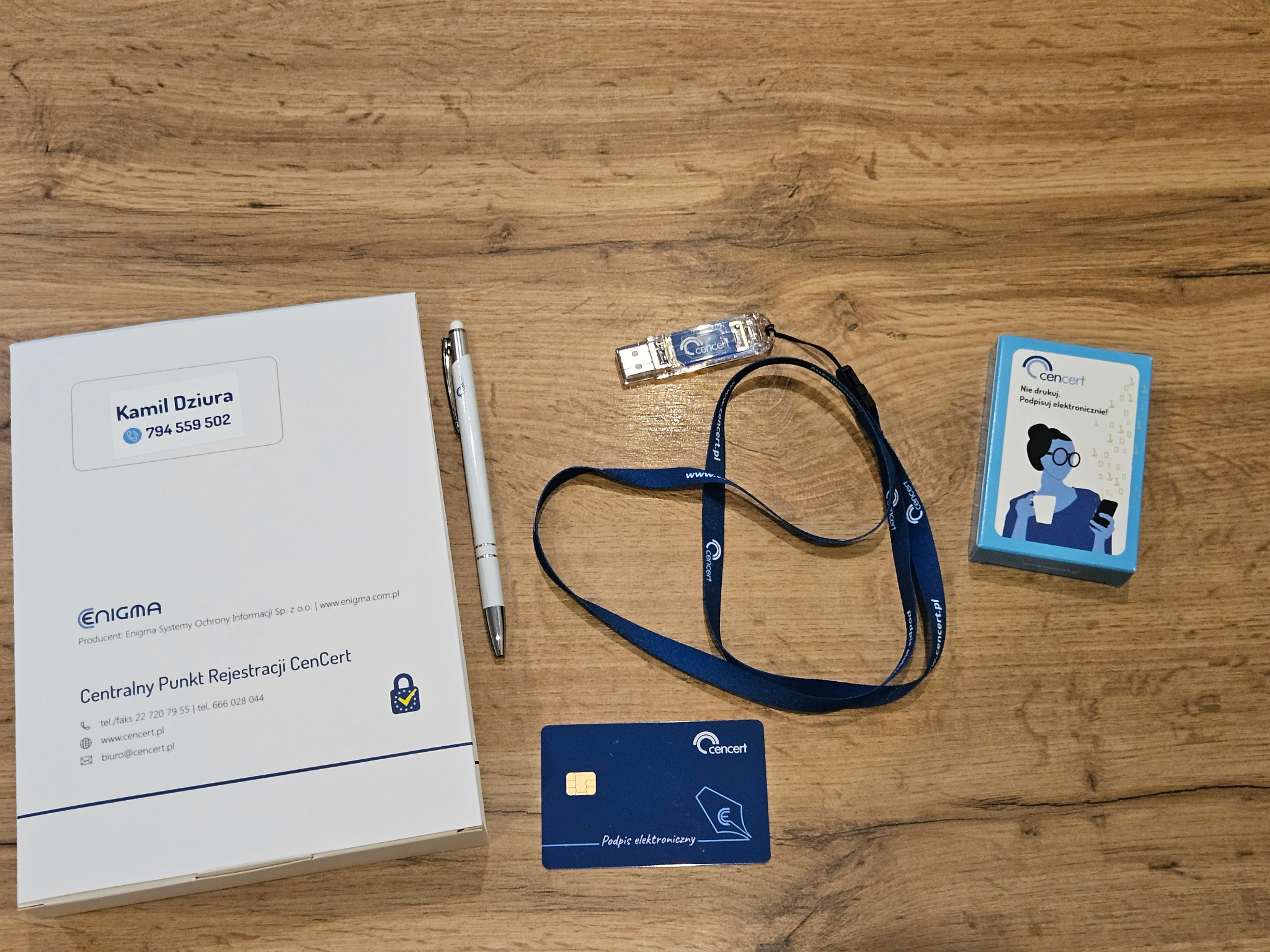 Zestaw materiałów informacyjnych dotyczących podpisu elektronicznego, zawierający folder z danymi kontaktowymi, długopis, kartę z chipem, smycz z logo firmy i pudełko z instrukcją, ułożone...