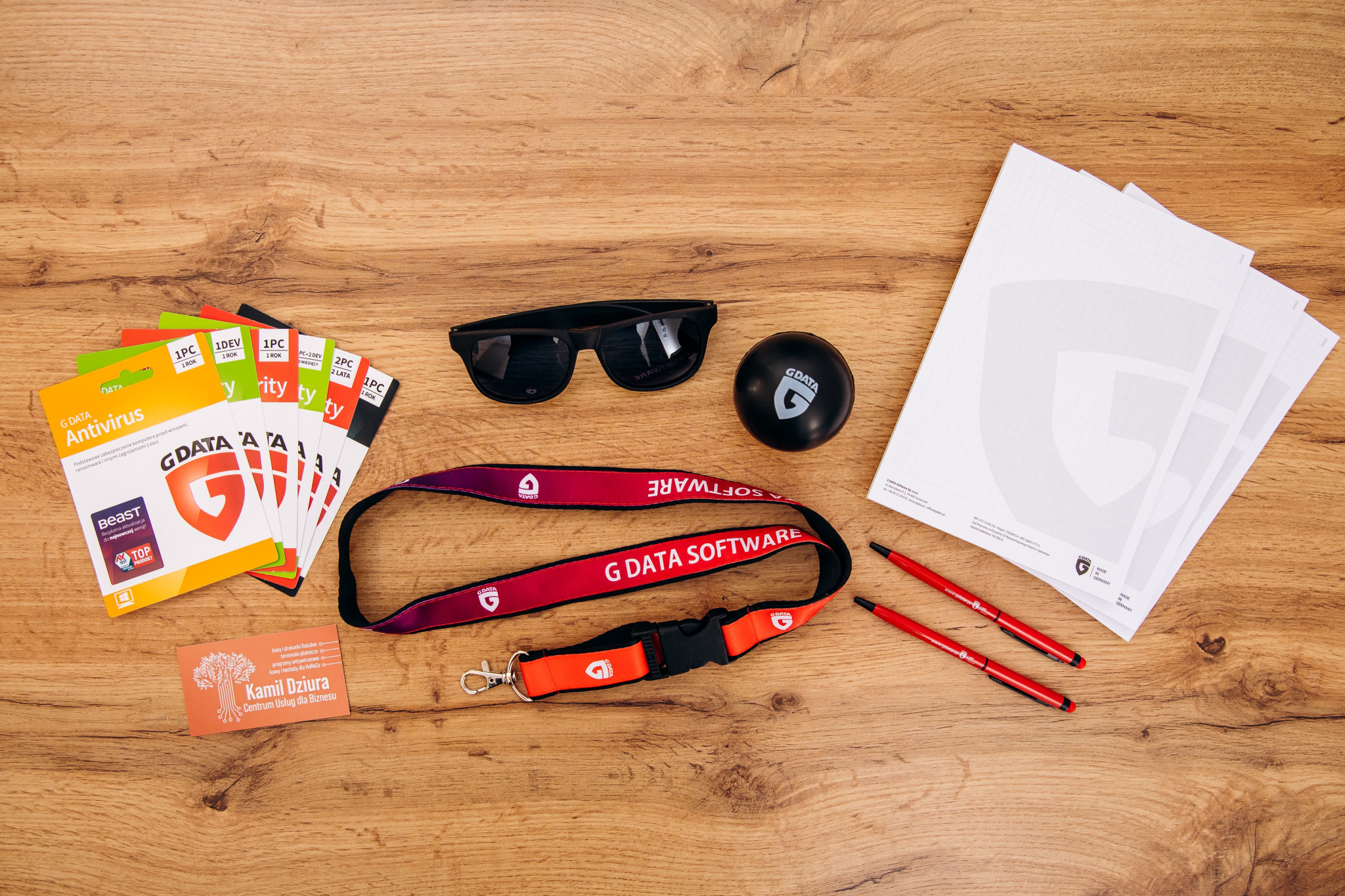 Biurko z drewnianym blatem, na którym leżą: opakowania programów antywirusowych G DATA, smycz G DATA SOFTWARE, piłeczka antystresowa G DATA, długopisy G DATA, wizytówka, okulary przeciwsłoneczne...