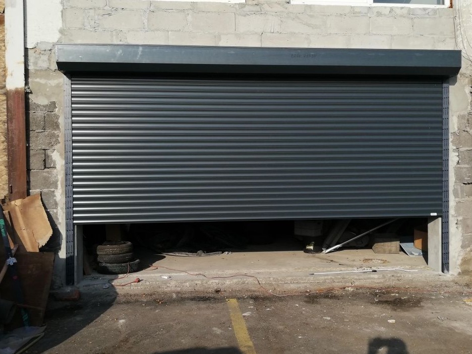 Częściowo opuszczona, szara roleta zewnętrzna garażowa z widocznymi oponami i narzędziami wewnątrz garażu, na tle surowego, szarego muru.