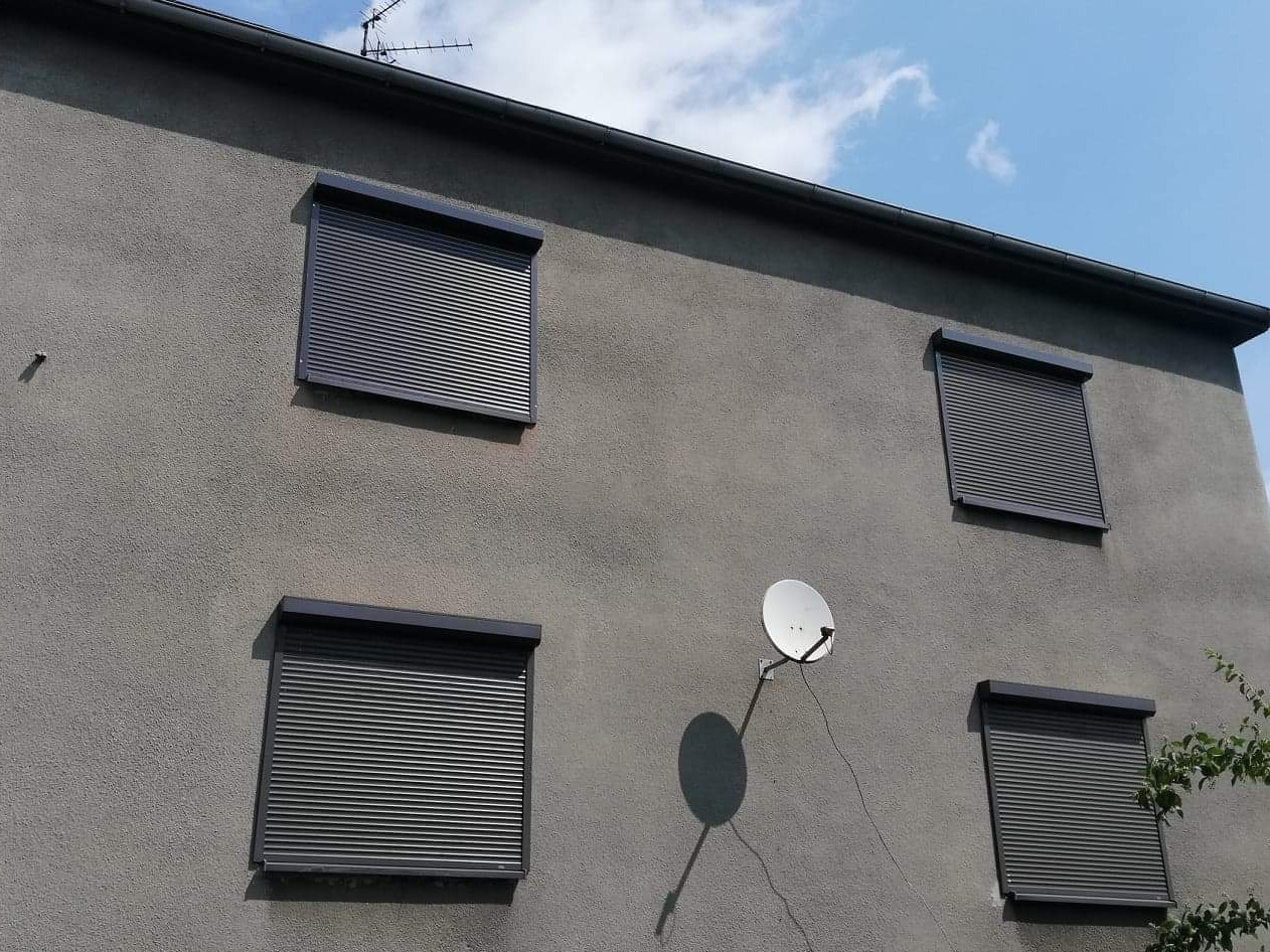 Elewacja budynku w Bytomiu z czterema oknami zabezpieczonymi szarymi roletami zewnętrznymi, antena satelitarna na ścianie, błękitne niebo w tle.