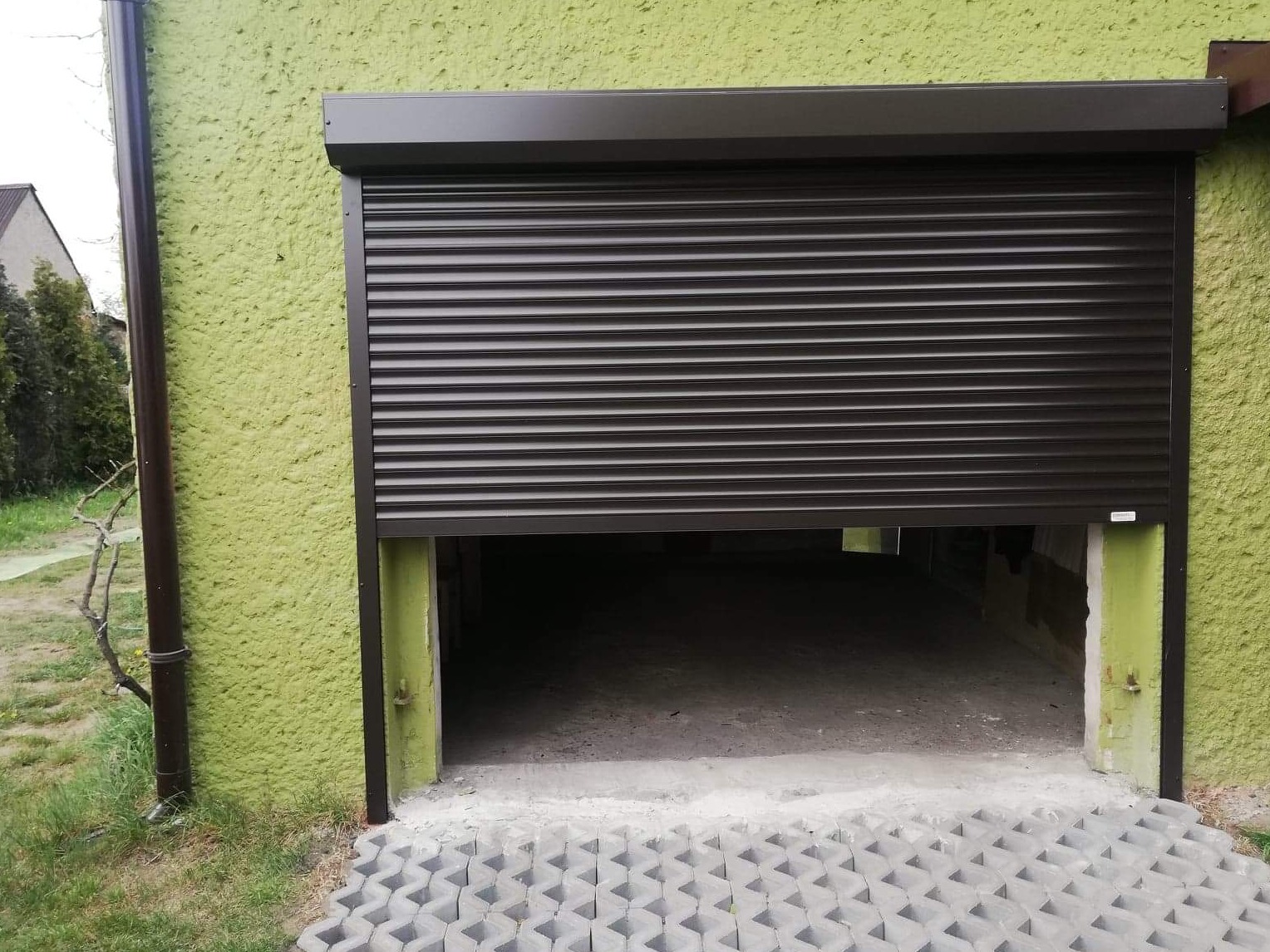 Brązowa brama garażowa rolowana, częściowo otwarta, na tle zielonej, chropowatej ściany budynku. Widoczne betonowe ażurowe płyty przed wjazdem i rynna spustowa po lewej stronie.