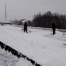 IMPLEX SERVICE GROUP Usługi Czystościowe - Dwóch pracowników usuwa śnieg z płaskiego dachu budynku przemysłowego w pochmurny dzień, widoczne urządzenia wentylacyjne i odśnieżone fragmenty dachu.