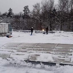 IMPLEX SERVICE GROUP Usługi Czystościowe - Pracownicy odśnieżają dach budynku przemysłowego, widoczne urządzenia wentylacyjne i świetliki pokryte śniegiem, zima.