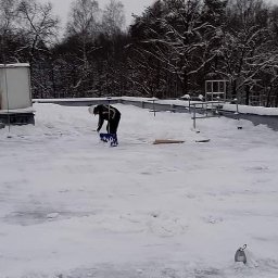 IMPLEX SERVICE GROUP Usługi Czystościowe - Pracownik w ciemnej kurtce usuwa śnieg z płaskiego dachu za pomocą niebieskiej łopaty. Na dachu widoczne urządzenia i warstwa śniegu, w tle zalesiony teren.