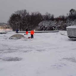 IMPLEX SERVICE GROUP Usługi Czystościowe - Pracownicy w pomarańczowych kamizelkach usuwają śnieg z płaskiego dachu budynku przemysłowego, na którym znajdują się duże agregaty klimatyzacyjne.