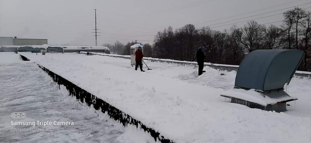 Dwóch pracowników usuwa śnieg z płaskiego dachu budynku przemysłowego w pochmurny dzień, widoczne urządzenia wentylacyjne i odśnieżone fragmenty dachu.