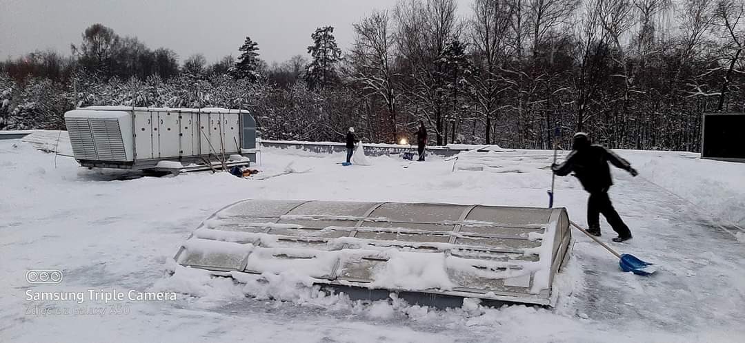 Pracownicy odśnieżają dach budynku przemysłowego, widoczne urządzenia wentylacyjne i świetliki pokryte śniegiem, zima.