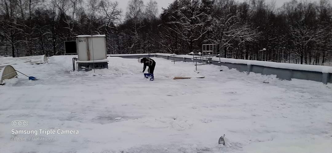 Pracownik w ciemnej kurtce usuwa śnieg z płaskiego dachu za pomocą niebieskiej łopaty. Na dachu widoczne urządzenia i warstwa śniegu, w tle zalesiony teren.