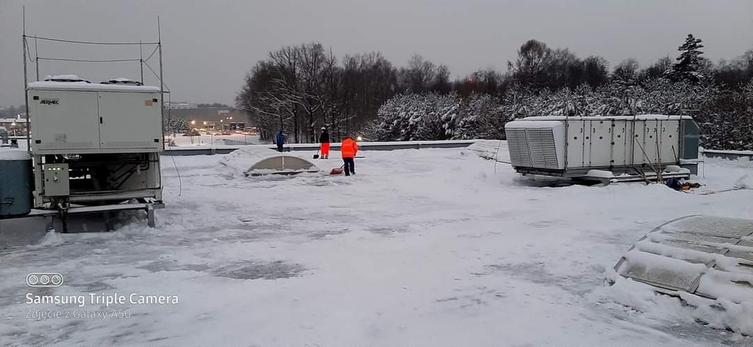Pracownicy w pomarańczowych kamizelkach usuwają śnieg z płaskiego dachu budynku przemysłowego, na którym znajdują się duże agregaty klimatyzacyjne.