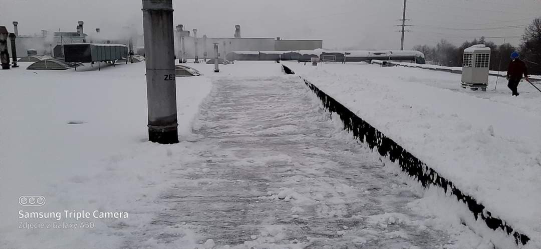 Śnieg na płaskim dachu budynku przemysłowego, częściowo odśnieżony chodnik, pracownik z łopatą w oddali, widoczne elementy wentylacji i kominy.