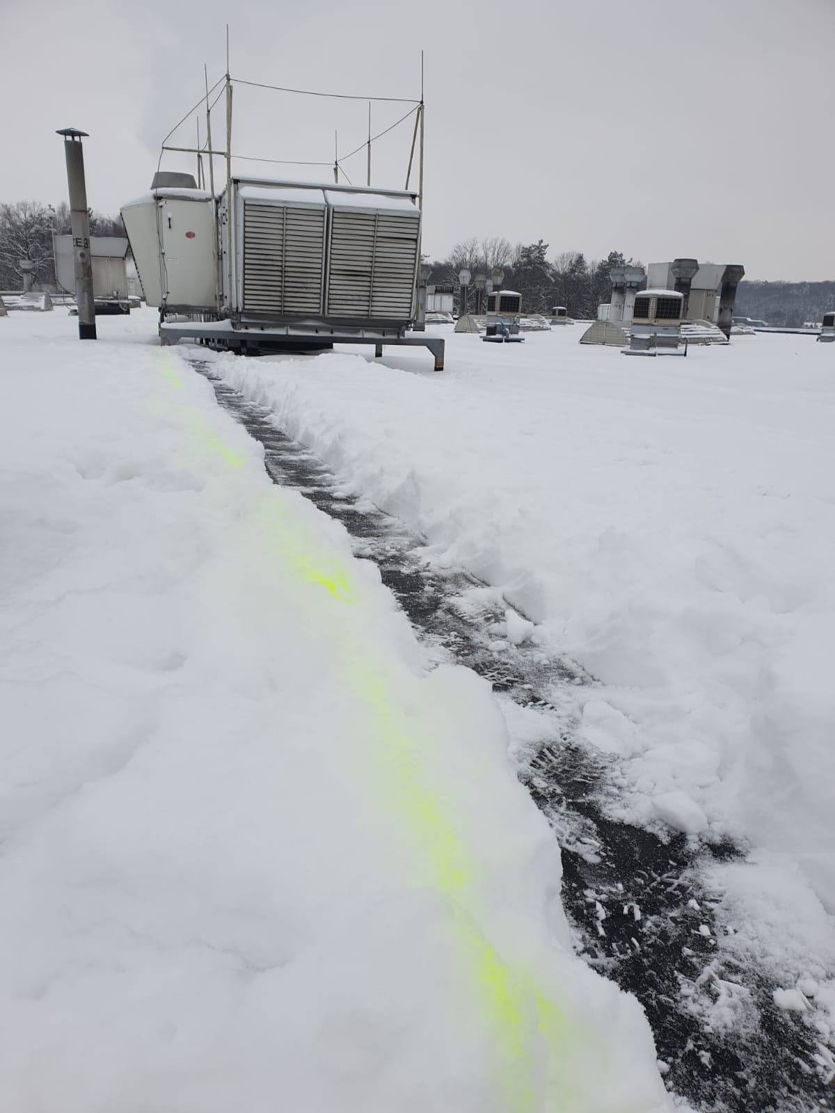 Śnieg usunięty z dachu budynku przemysłowego, odsłaniając czarną powierzchnię; w tle widoczne instalacje wentylacyjne i drzewa bez liści pod szarym niebem.