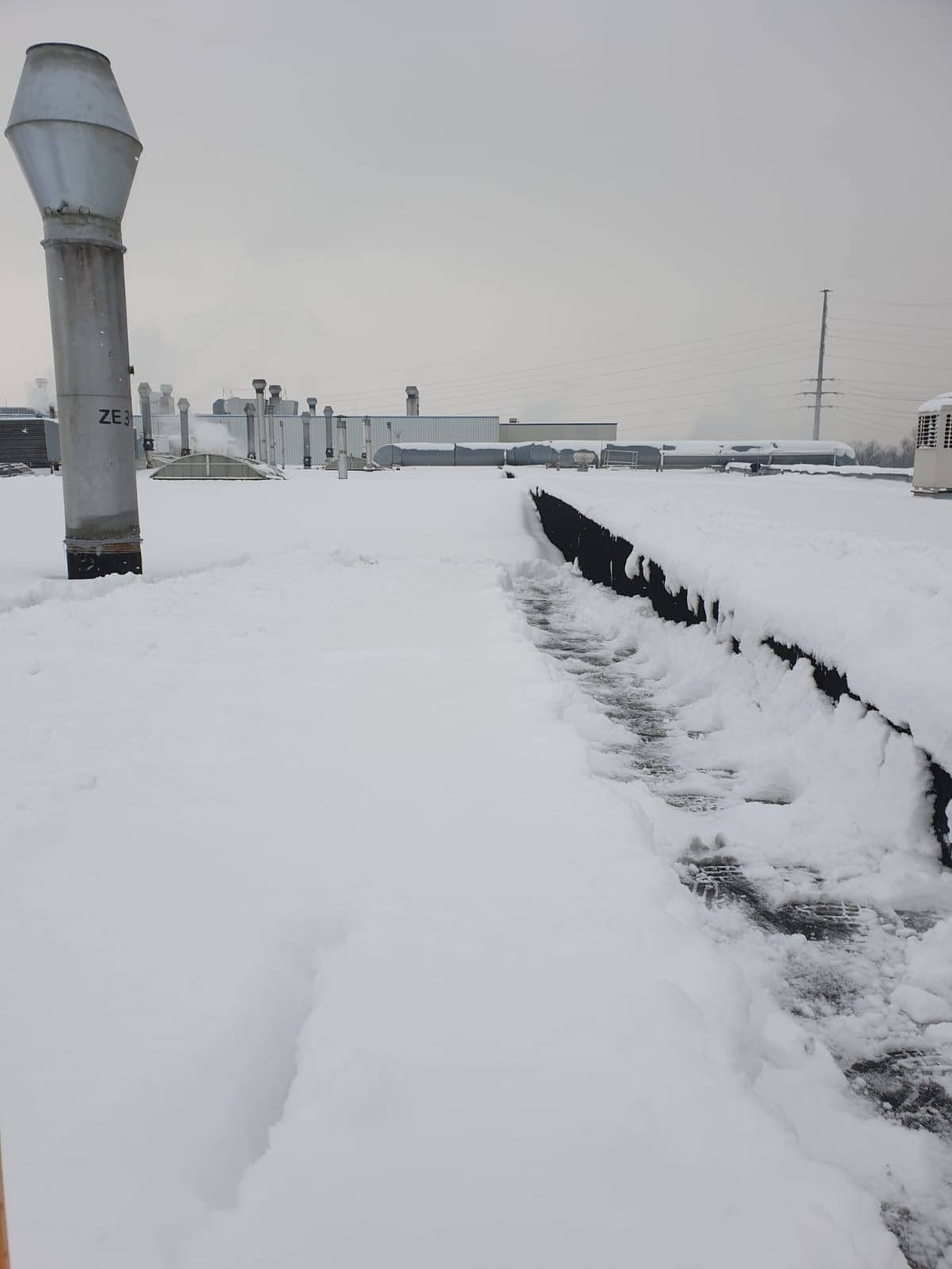 Śnieżny dach przemysłowy z odśnieżoną ścieżką, widoczne kominy wentylacyjne i instalacje techniczne w tle, pochmurne niebo.