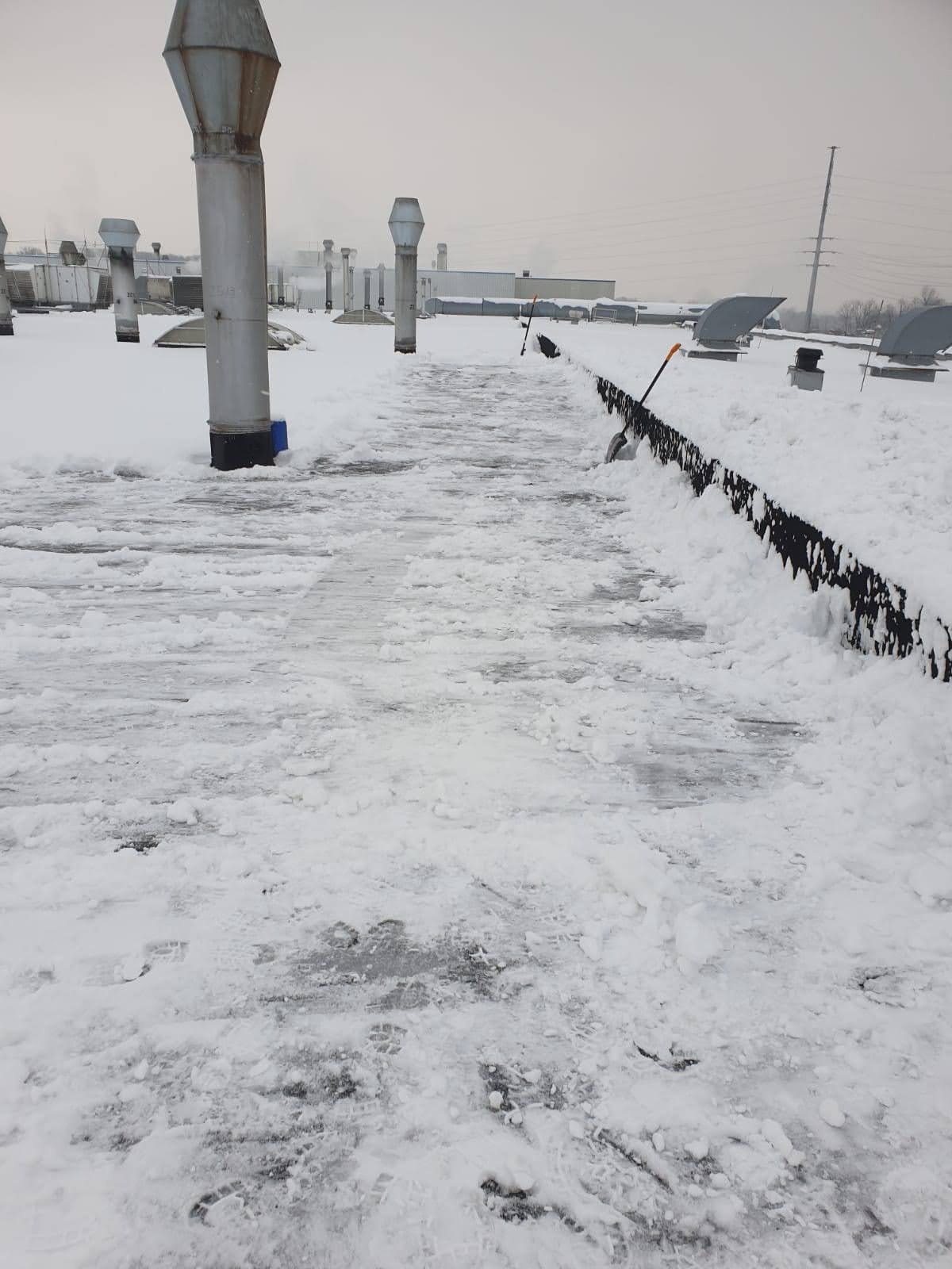 Odśnieżony fragment dachu przemysłowego z widocznymi kominami wentylacyjnymi, resztki śniegu i narzędzia do odśnieżania przy krawędzi dachu.