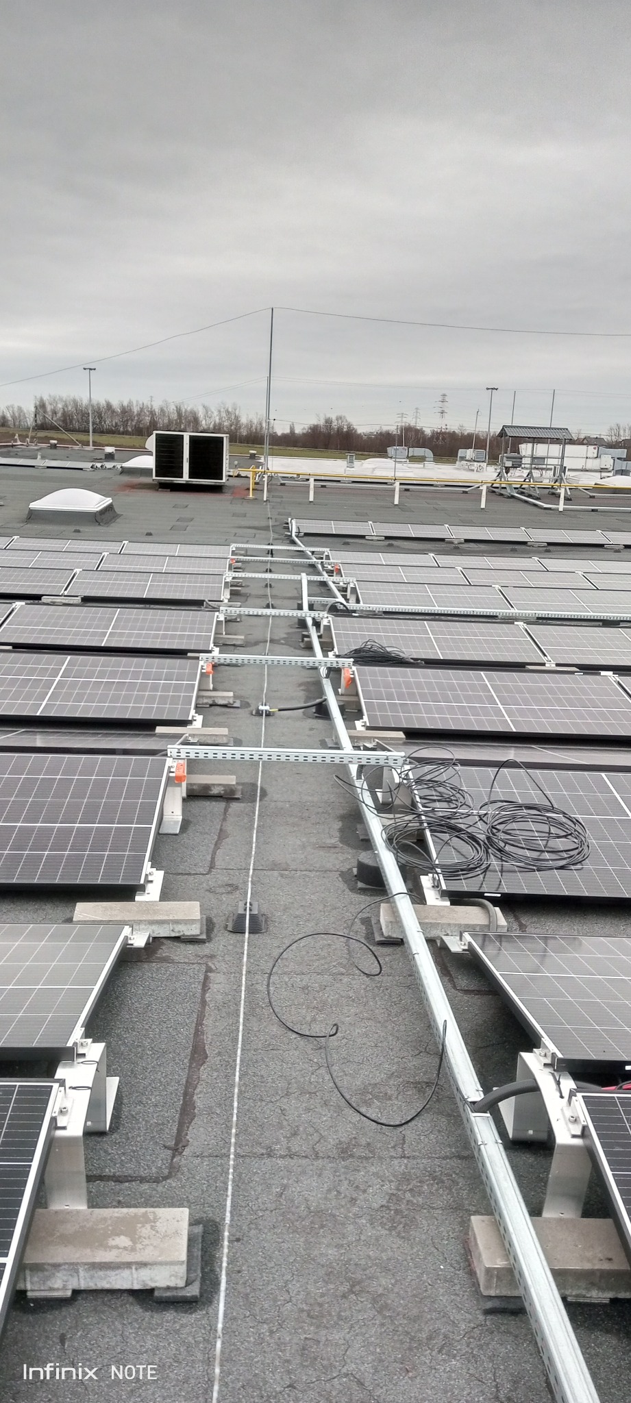 Widok z góry na instalację paneli słonecznych na płaskim dachu, z widocznymi elementami konstrukcyjnymi i okablowaniem, na tle pochmurnego nieba.