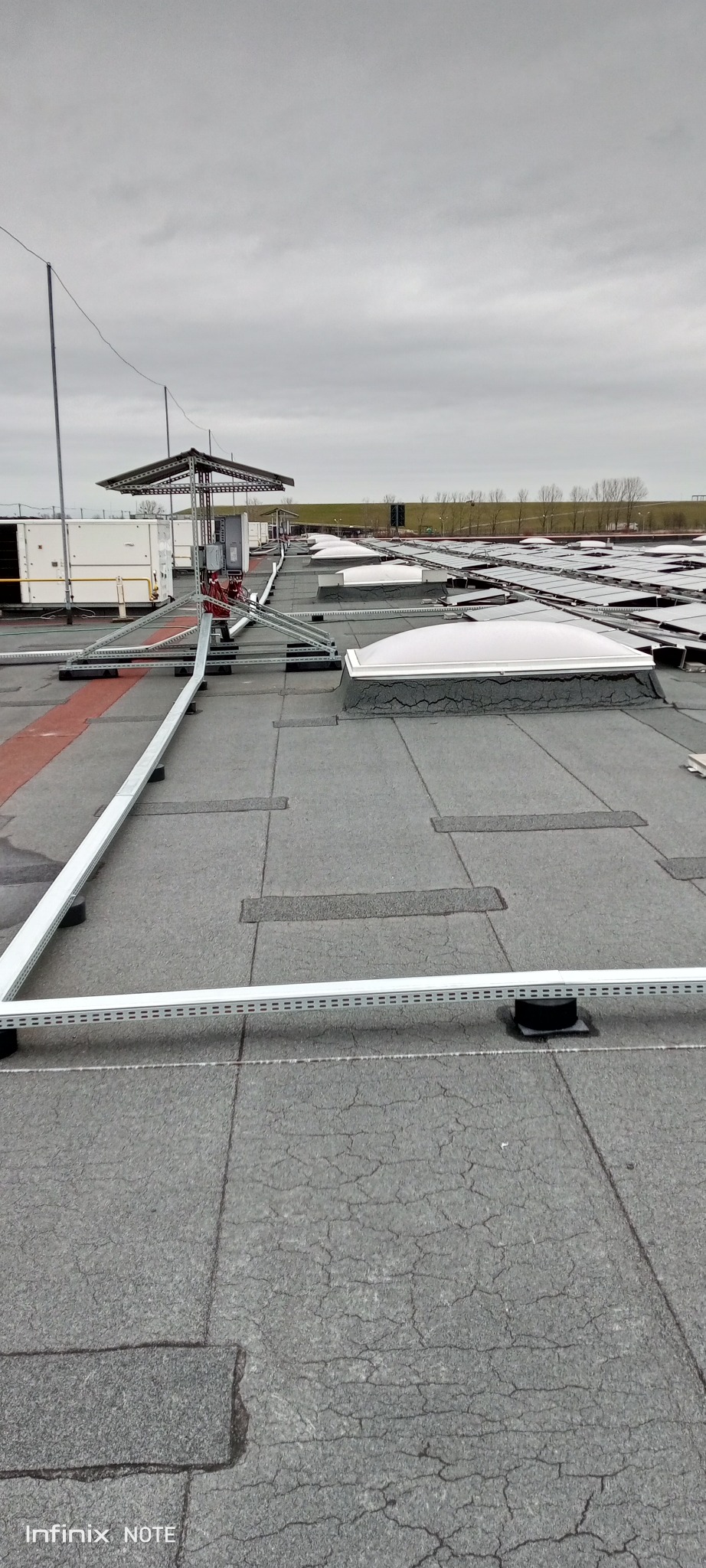 Rozległy, płaski dach pokryty papą z widocznymi świetlikami dachowymi, systemem odgromowym i instalacjami technicznymi w pochmurny dzień.