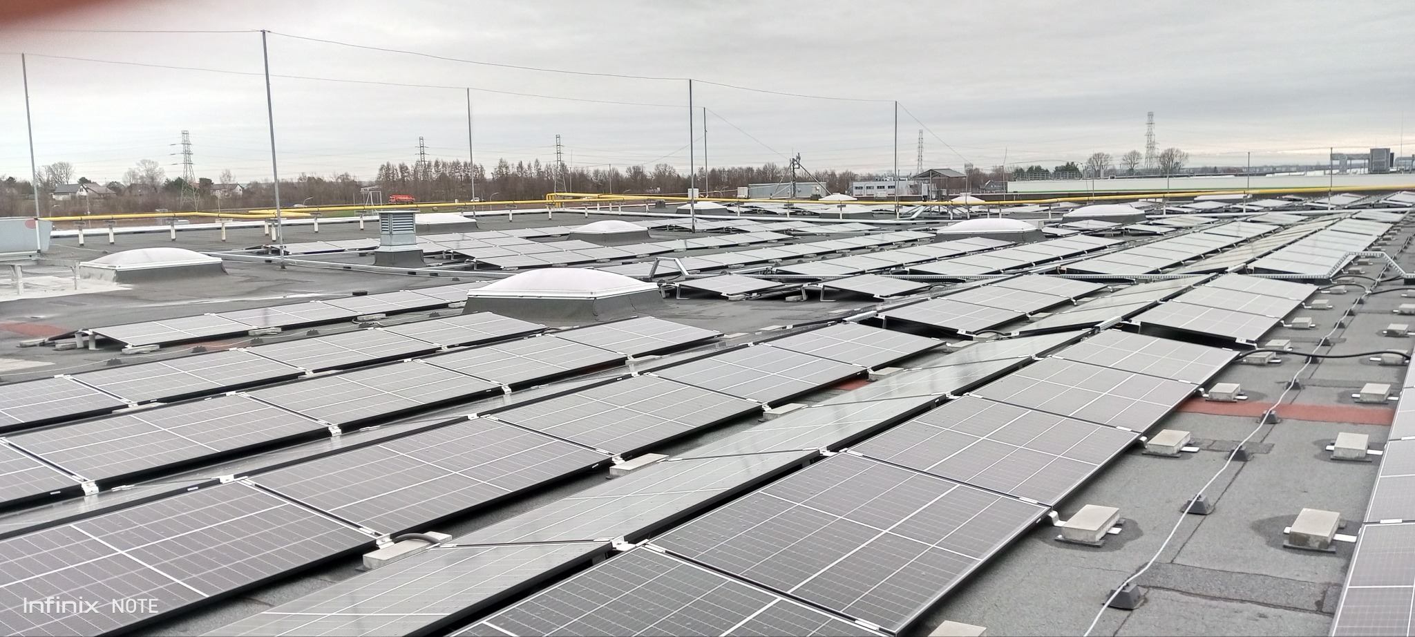 Widok z góry na płaski dach pokryty panelami słonecznymi, z widocznymi świetlikami dachowymi i elementami instalacji odgromowej na tle pochmurnego nieba i zabudowań.