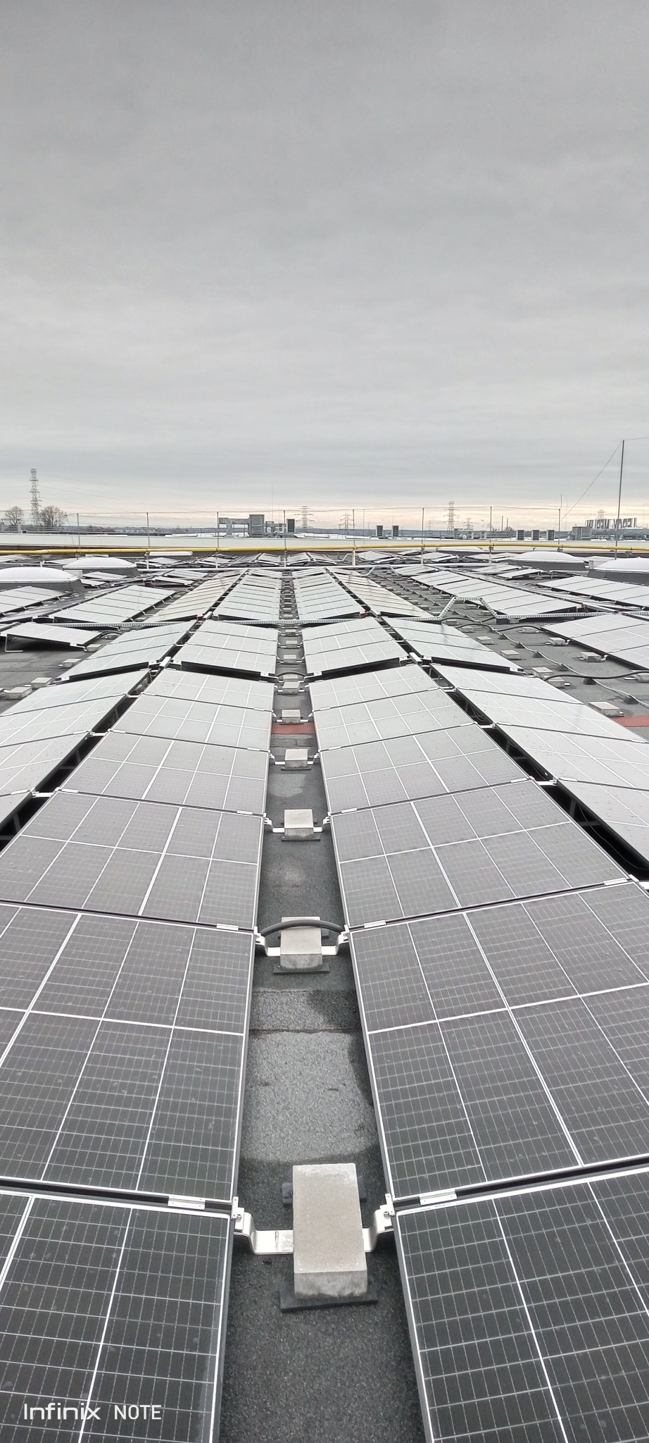 Widok z góry na dużą instalację paneli słonecznych na dachu budynku w pochmurny dzień. Widoczne mocowania i kable. W tle zarys zabudowań i słupy wysokiego napięcia.