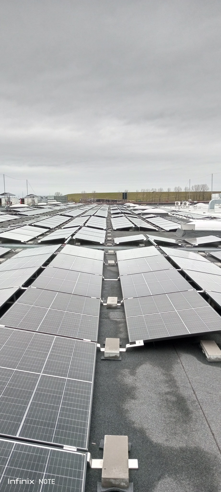 Rozległy widok z góry na dach pokryty panelami słonecznymi w pochmurny dzień, z widocznymi elementami mocującymi i odległym horyzontem.