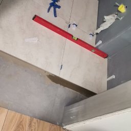 M-Bud - Układanie beżowych płytek ściennych z użyciem poziomicy i plastikowych krzyżyków dystansowych, widoczna podłoga wyłożona panelami drewnopodobnymi oraz wiadro z zaprawą.