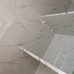 M-Bud - Ściana wyłożona białymi płytkami imitującymi marmur z zamontowanymi półkami, widoczne białe wsporniki półek, ujęcie z dołu.