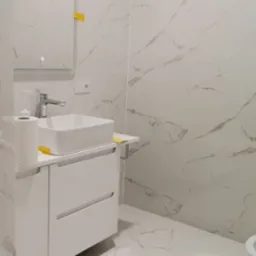 Nowoczesna łazienka z białymi płytkami imitującymi marmur na ścianach i podłodze, biała szafka pod umywalką z prostokątną umywalką nablatową, lustro z podświetleniem LED, fragment toalety w tle.