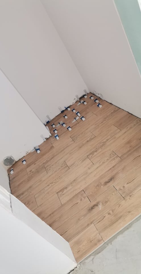 Układanie podłogi z płytek imitujących drewno w rogu pomieszczenia, z użyciem niebieskich krzyżyków dystansowych, widoczny fragment ściany i rura wentylacyjna.