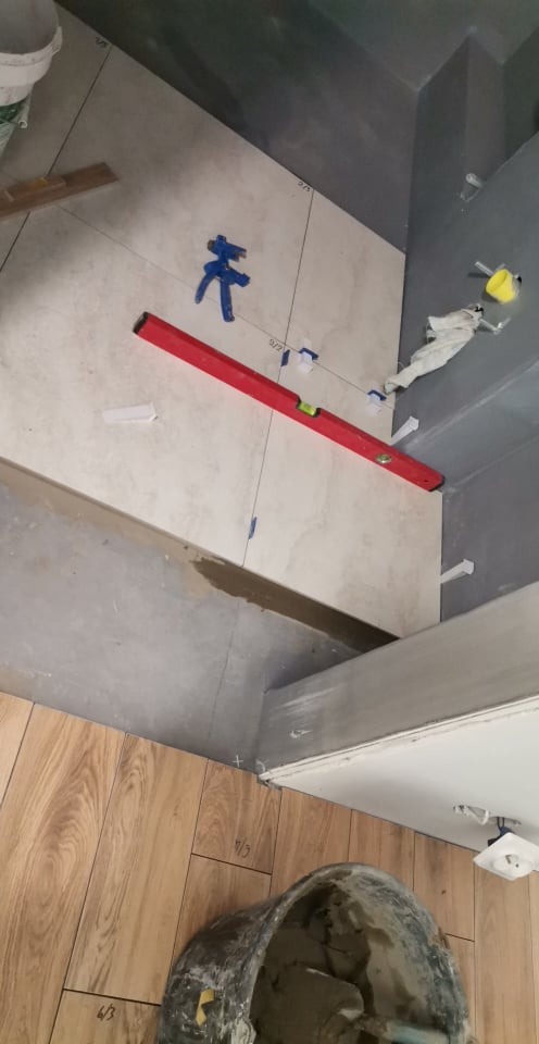 Układanie beżowych płytek ściennych z użyciem poziomicy i plastikowych krzyżyków dystansowych, widoczna podłoga wyłożona panelami drewnopodobnymi oraz wiadro z zaprawą.