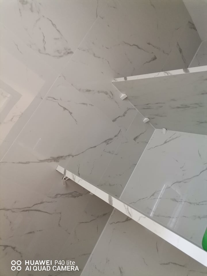 Ściana wyłożona białymi płytkami imitującymi marmur z zamontowanymi półkami, widoczne białe wsporniki półek, ujęcie z dołu.