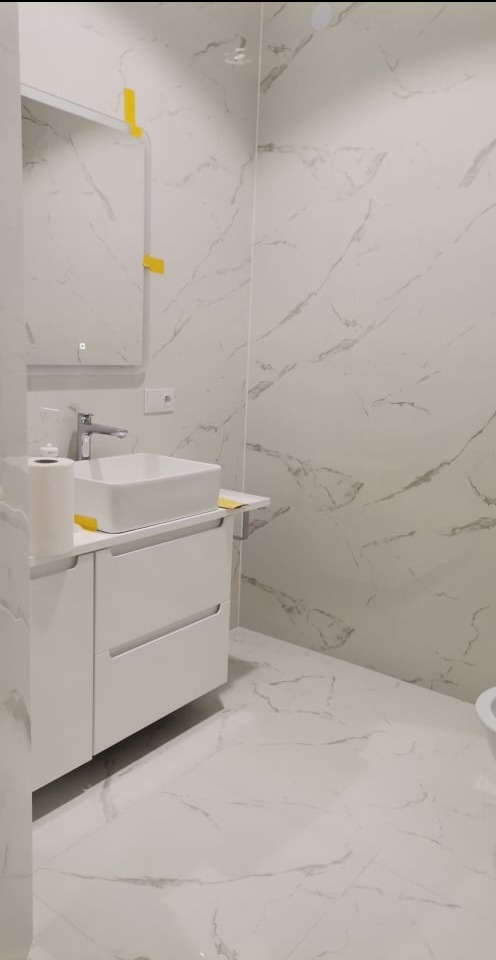 Nowoczesna łazienka z białymi płytkami imitującymi marmur na ścianach i podłodze, biała szafka pod umywalką z prostokątną umywalką nablatową, lustro z podświetleniem LED, fragment toalety w tle.