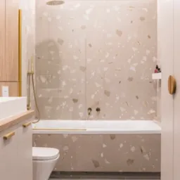 Nowoczesna łazienka z beżowymi płytkami w terrazzo, złotą armaturą i drewnianymi akcentami, widok z perspektywy wejścia.