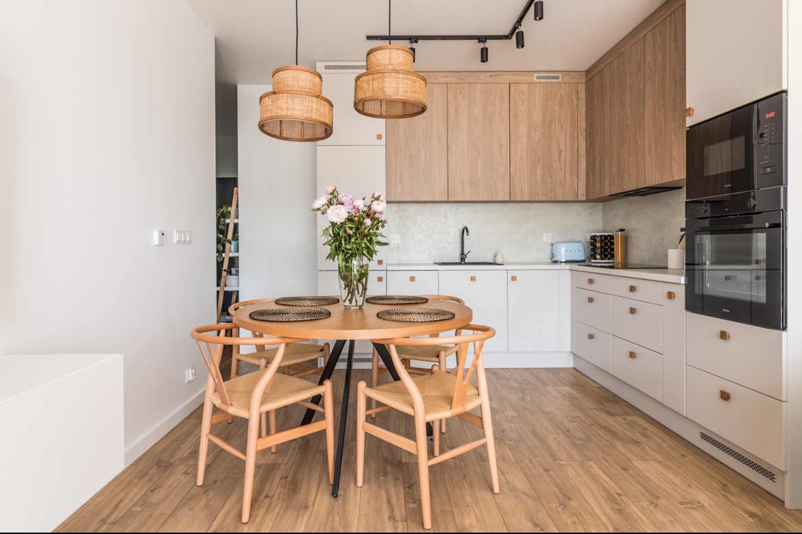 Jasna kuchnia z drewnianymi akcentami: okrągły stół z kwiatami, rattanowe lampy, zabudowa kuchenna z piekarnikiem i mikrofalówką.