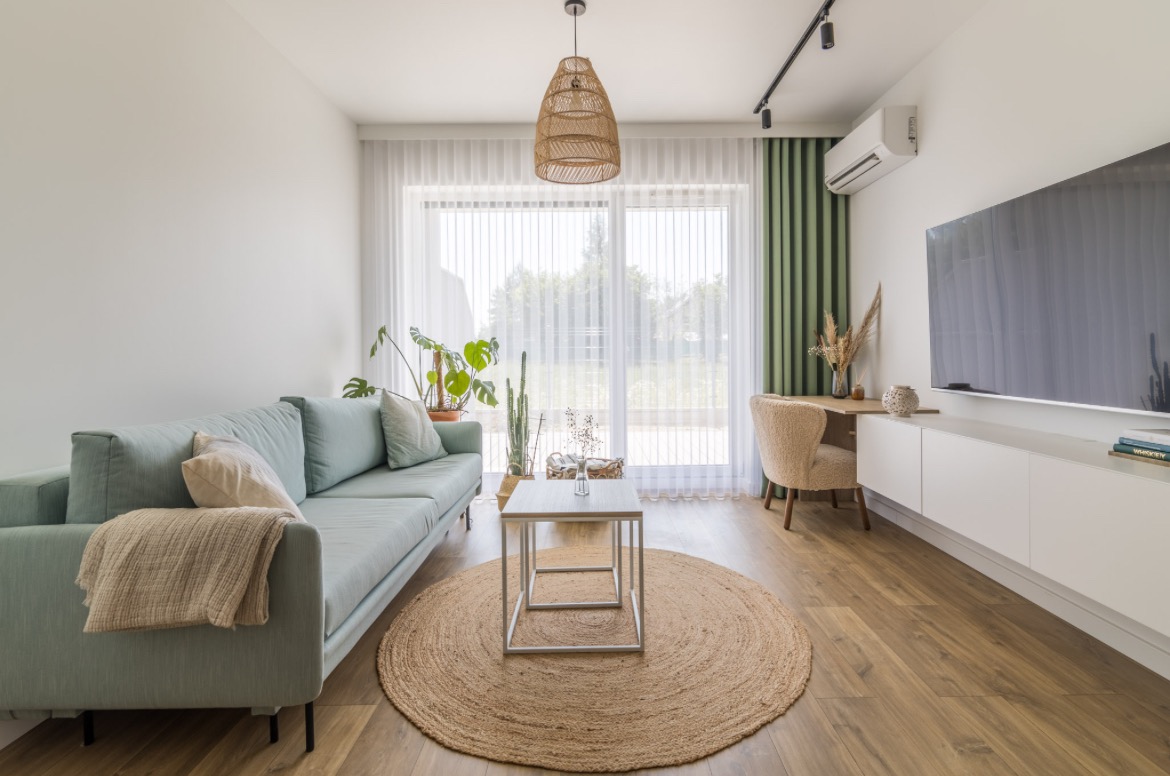 Jasny salon z turkusową sofą, okrągłym jutowym dywanem i minimalistyczną szafką RTV pod telewizorem, oświetlony wiszącą lampą z naturalnego materiału.