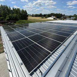 MICHAŁ KOWALSKI ELEKTROMAX - Widok z góry na zainstalowane panele fotowoltaiczne na metalowym dachu budynku. W tle drzewa i błękitne niebo z chmurami.