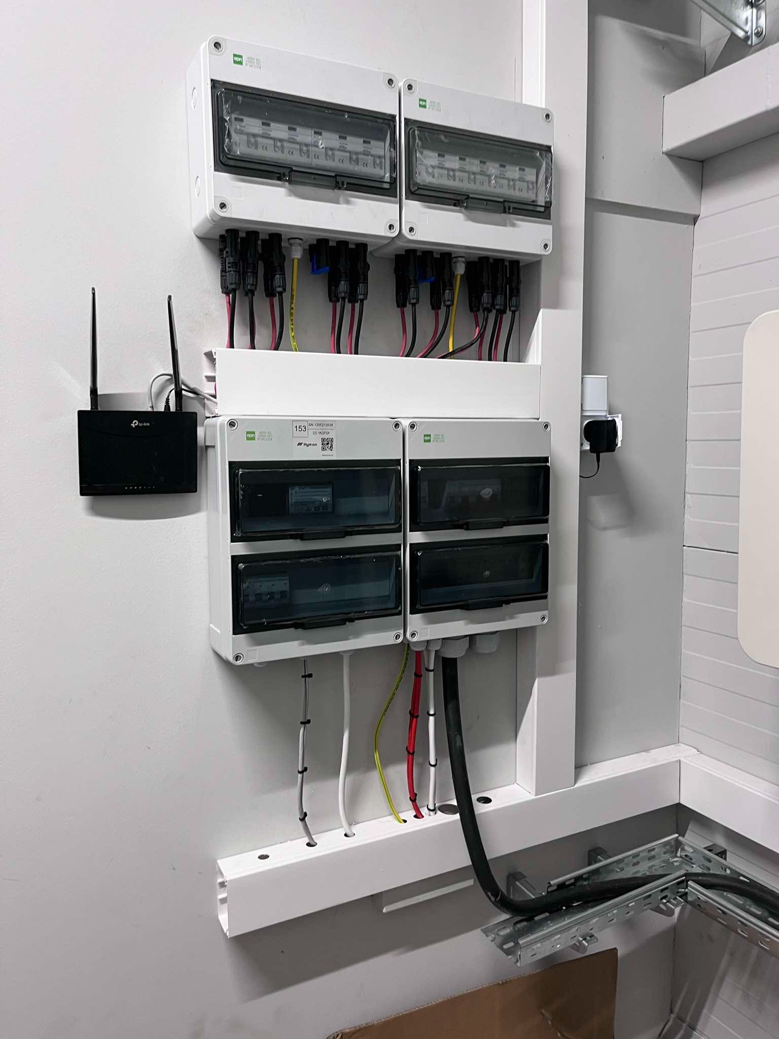 Rozdzielnica elektryczna z podłączonymi przewodami i routerem TP-Link na ścianie. Widoczne zabezpieczenia i kable w korytku. Całość w jasnej kolorystyce.