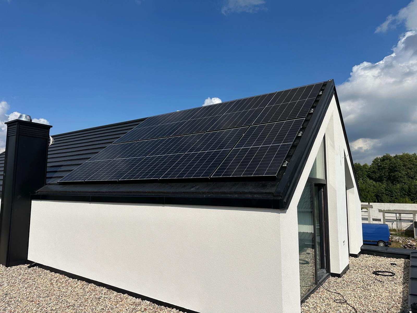 Nowoczesny dom z panelami fotowoltaicznymi na dachu w słoneczny dzień. Biała elewacja kontrastuje z czarną dachówką i panelami. Widoczny komin i okno.