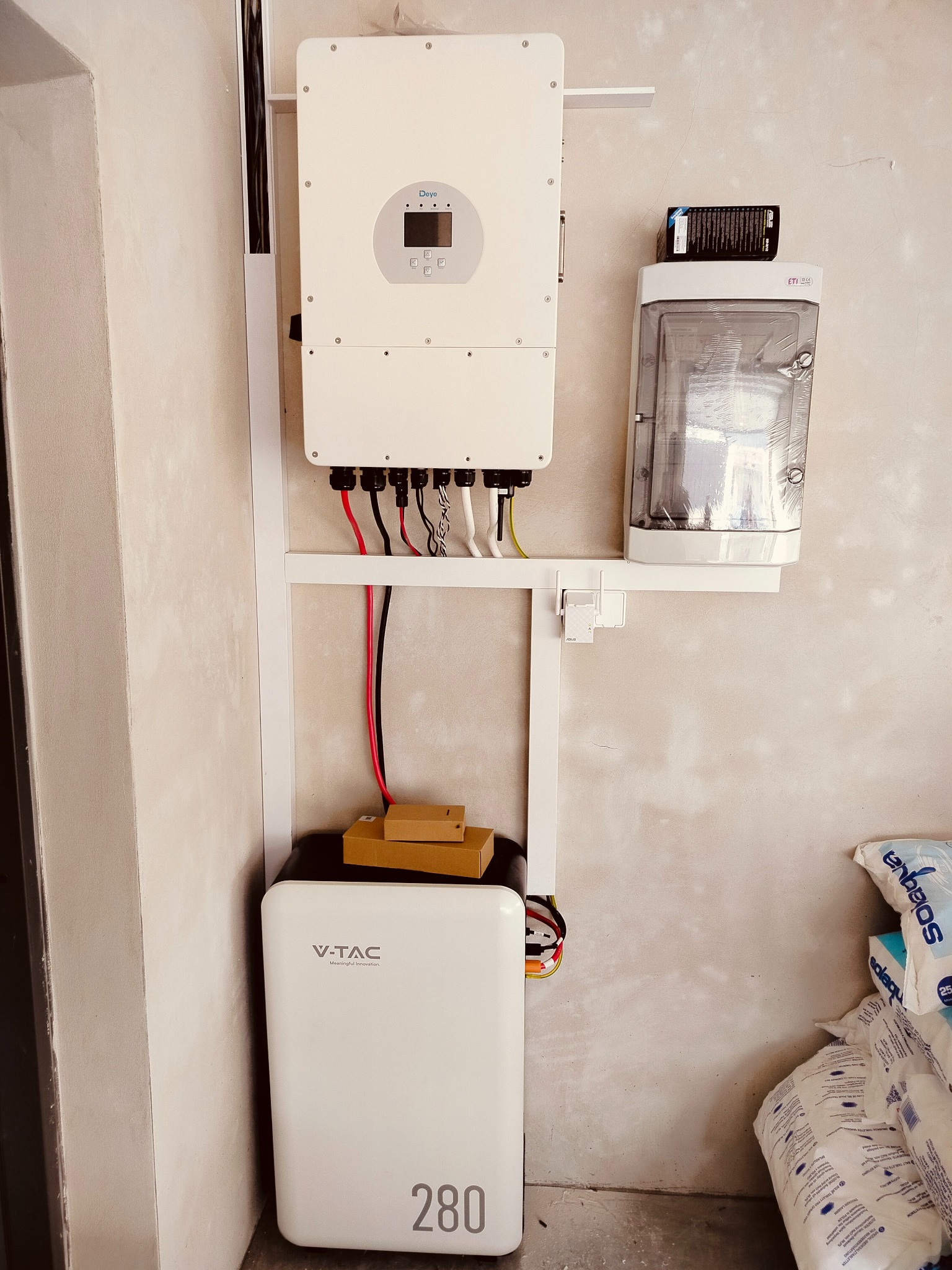 Biały inwerter Deye, akumulator V-TAC 280 i skrzynka ETI zamontowane na ścianie na białym stelażu. Widoczne kable. Obok leżą worki z solą.