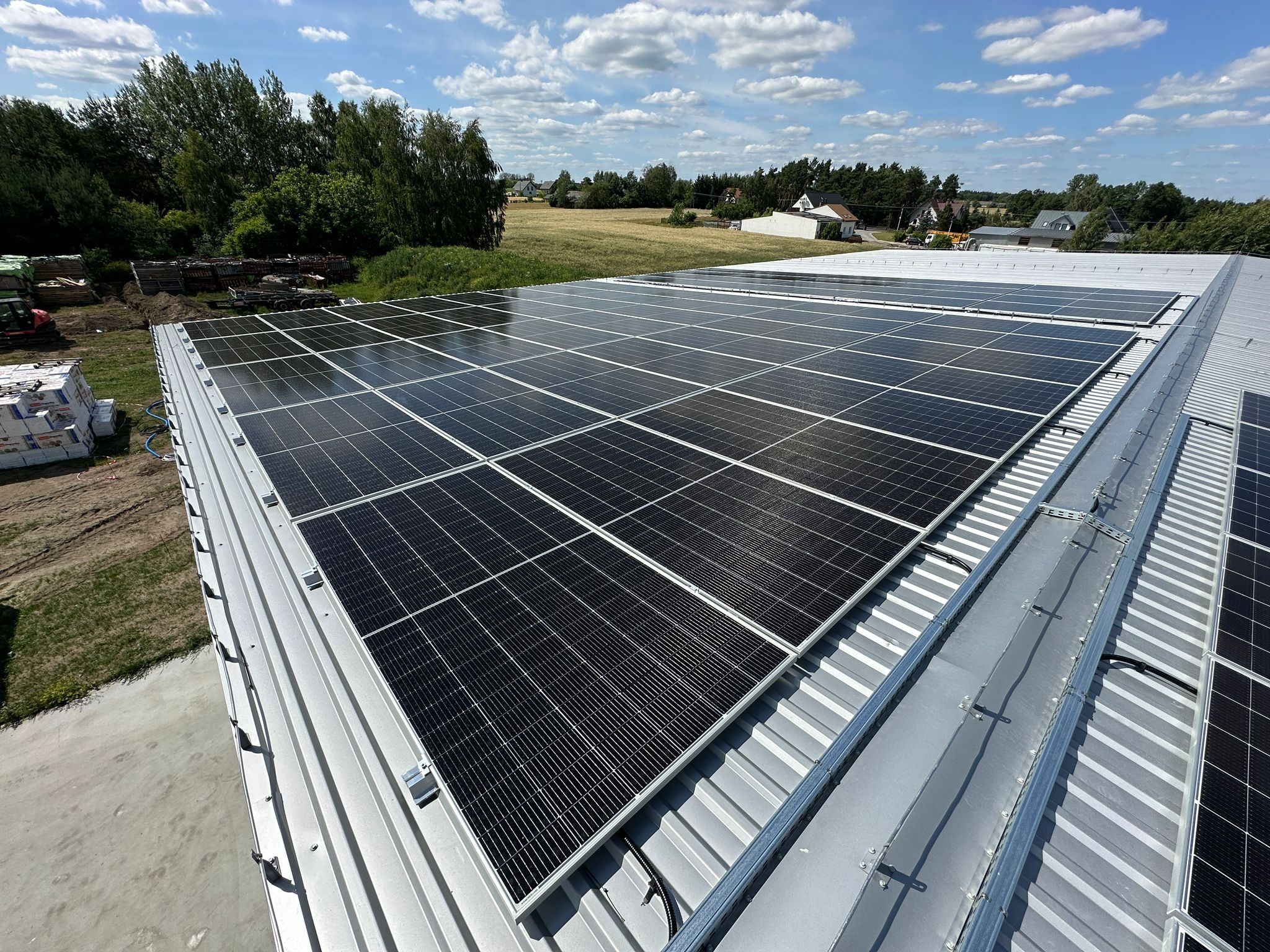 Widok z góry na zainstalowane panele fotowoltaiczne na metalowym dachu budynku. W tle drzewa i błękitne niebo z chmurami.