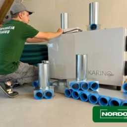 Montaż centrali wentylacyjnej z rekuperacją marki Karino przez instalatora w zielonej koszulce z napisem 'Rekuperacja na Śląsku'. Widoczne elementy instalacji, rury wentylacyjne w tle i logo Nordom...