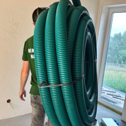 Adrian Dworak P.H.U. Nordom - Instalator niesie zwój zielonych, karbowanych rur wentylacyjnych na ramieniu w surowym wnętrzu budynku, obok widoczne kanały wentylacyjne i okno.