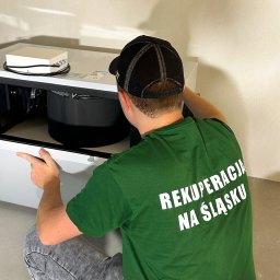 Adrian Dworak P.H.U. Nordom - Instalator w zielonej koszulce z napisem 'Rekuperacja na Śląsku' kuca przed białą jednostką rekuperacyjną podczas montażu, widok z tyłu, na ścianie widoczny napis '77'.
