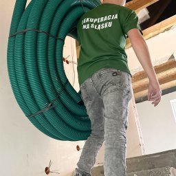 Adrian Dworak P.H.U. Nordom - Instalator w zielonej koszulce z napisem 'Rekuperacja na Śląsku' niesie zwój zielonych rur wentylacyjnych po betonowych schodach w trakcie montażu systemu rekuperacji w budynku w stanie surowym.