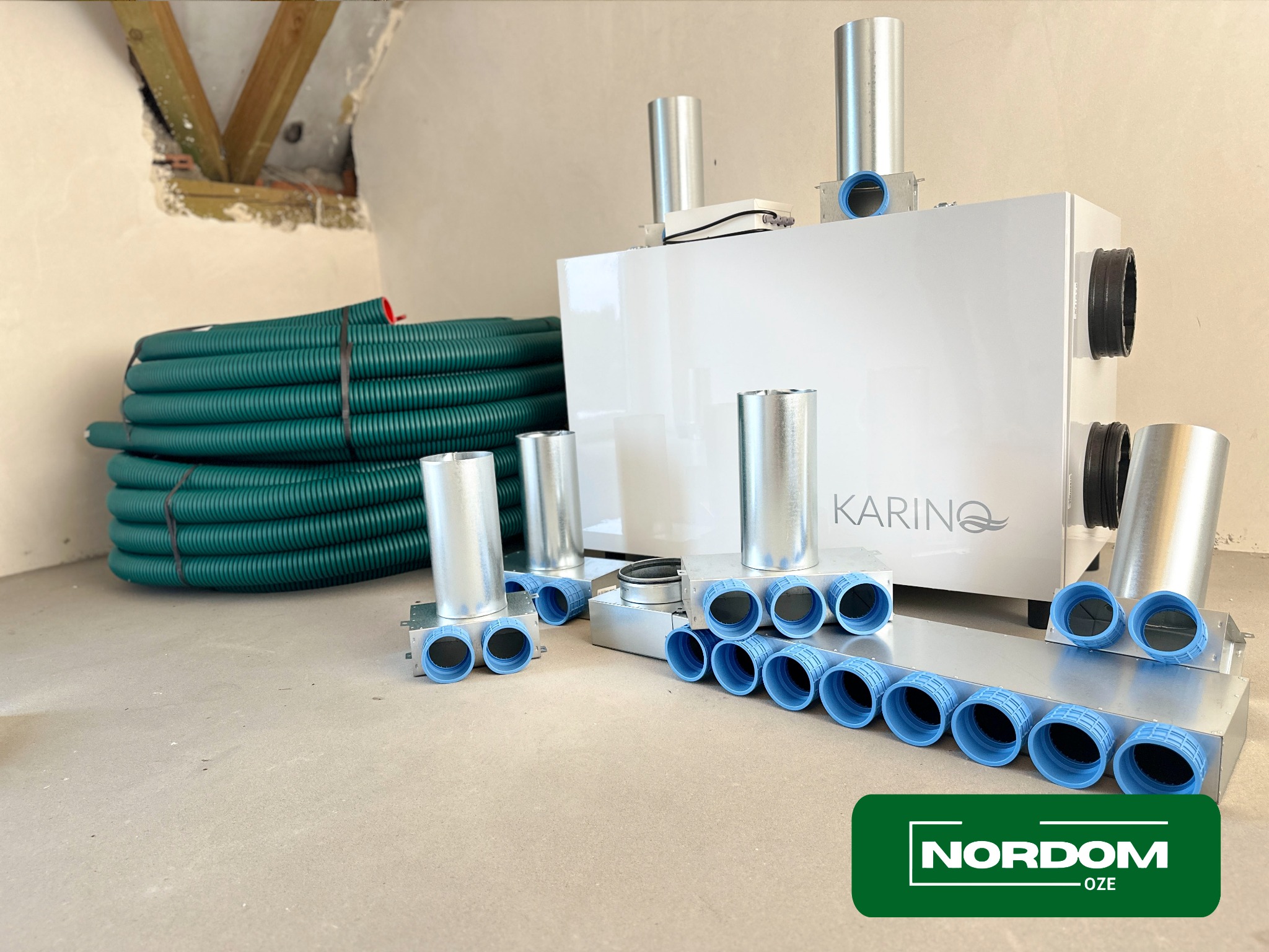 Biała centrala wentylacyjna z logo Karino, zielone rury wentylacyjne i metalowe rozdzielacze z niebieskimi uszczelkami na surowej podłodze w trakcie montażu, widoczny fragment ściany w stanie...